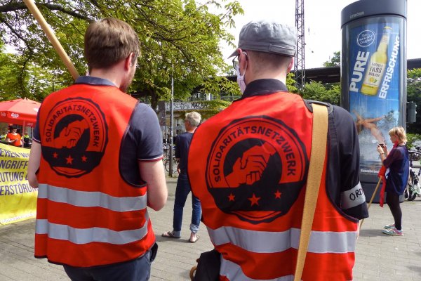 Man sieht zwei Männer von hinten, die die roten Westen vom Solidaritätsnetzwerk Deutschland tragen